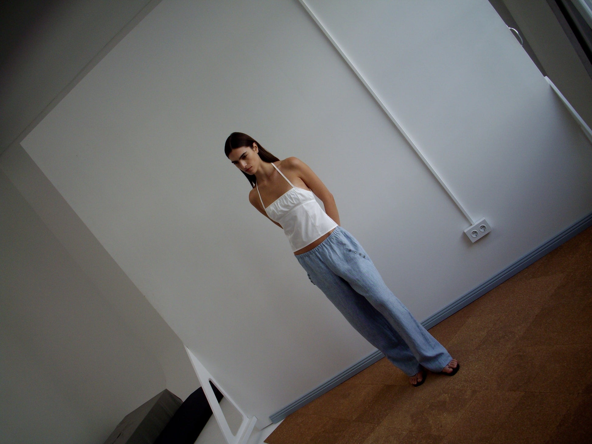 A woman wears the Strap Top - White by Deiji Studios with loose blue pants, standing against a plain white wall in a minimalist room, gazing slightly downward with her hands behind her back.