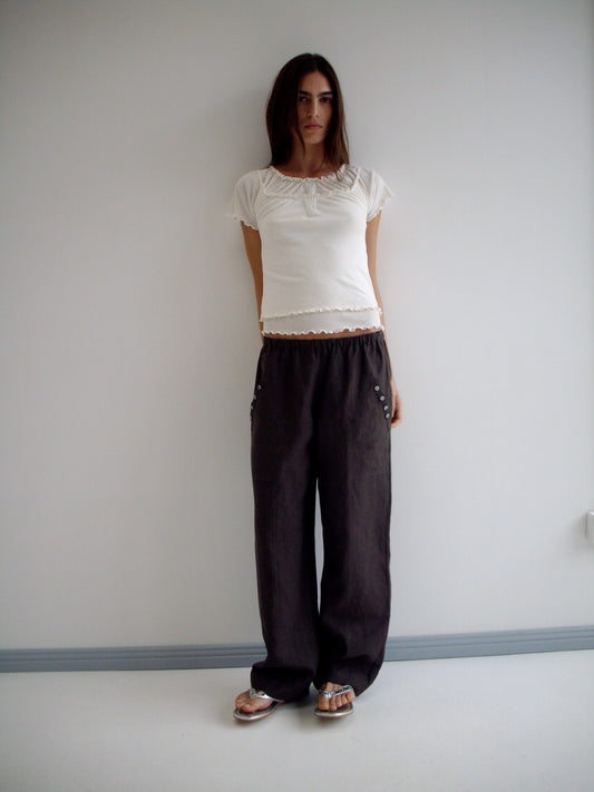 A woman with long dark hair and a neutral expression stands against a plain white wall wearing the Flounce Tee - Cream by Deiji Studios, paired with loose dark pants and sandals.