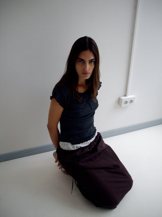 A woman with long brown hair kneels on a white floor in a minimalist room, wearing the Flounce Tee - Grey and a brown skirt. She looks directly at the camera with a serious expression.