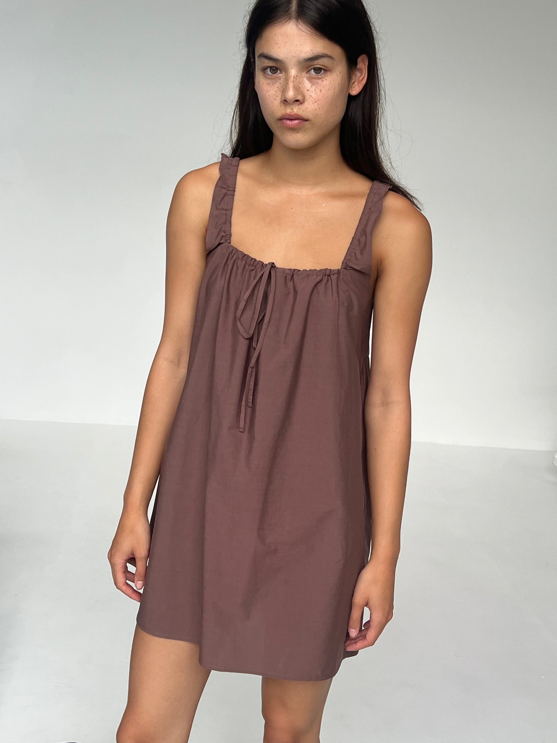 A woman with long dark hair wears The Bow Dress - Cedar by Deiji Studios, a sleeveless, above-the-knee dress in mauve organic cotton with ruffled straps and a neckline tie, standing against a plain white background.