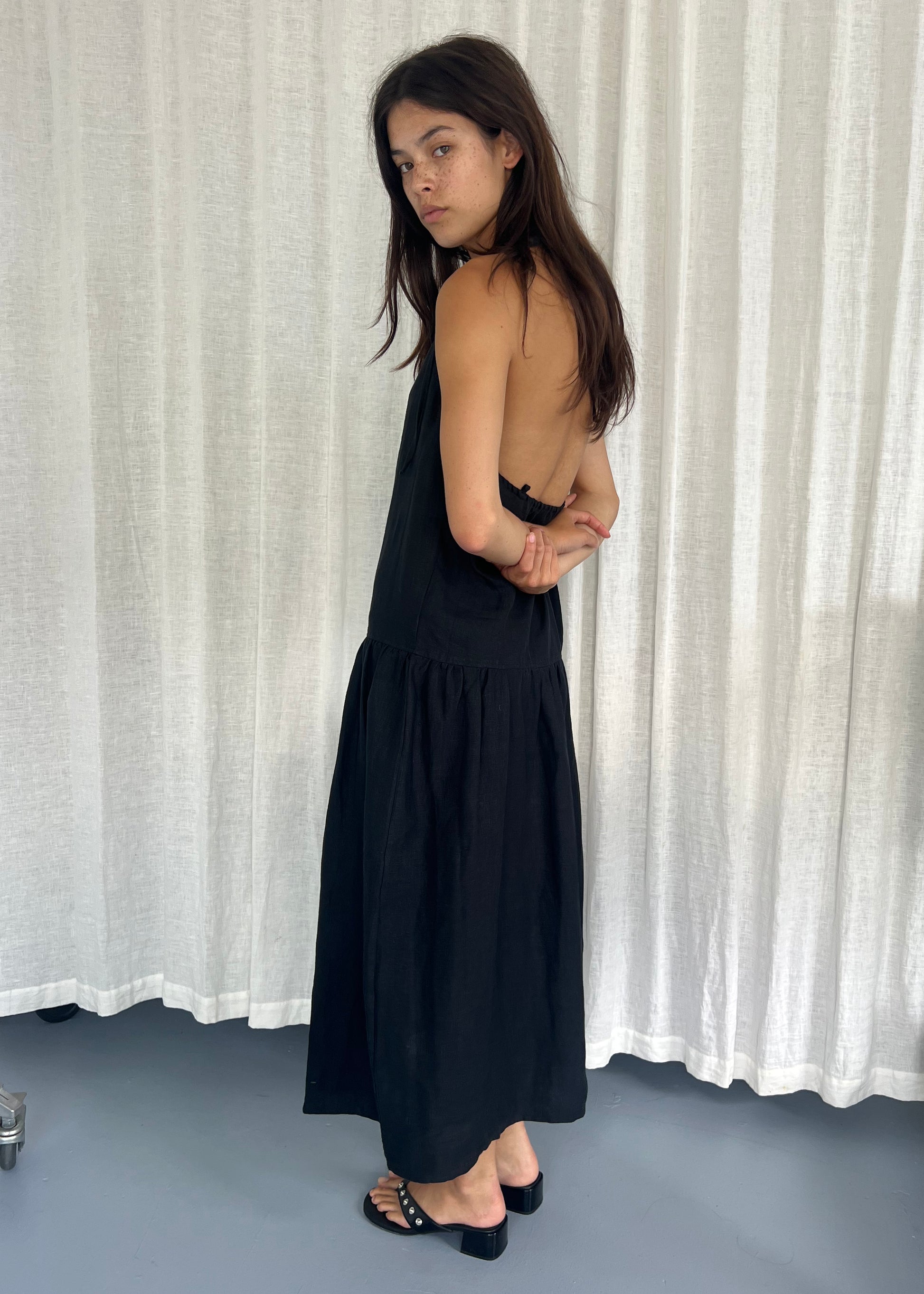 A woman with long dark hair stands sideways in front of white curtains, wearing the Gathered Halter Dress - Black and black sandals. She has her arms behind her back and looks over her shoulder at the camera.
