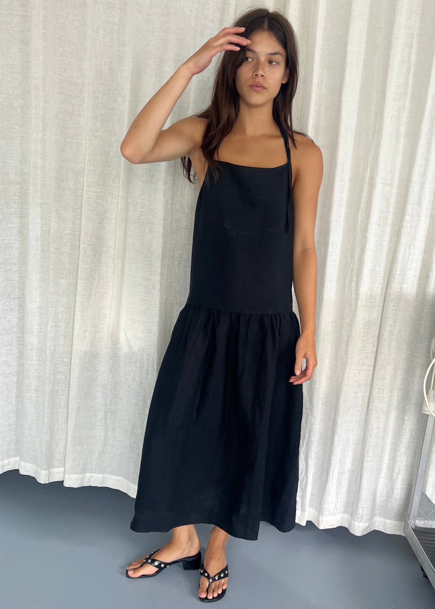 A woman stands indoors before white curtains, wearing the Gathered Halter Dress - Black by Deiji Studios. The sleeveless dress features a square neckline and ruffled hem. She is barefoot, touching her forehead with a neutral expression.