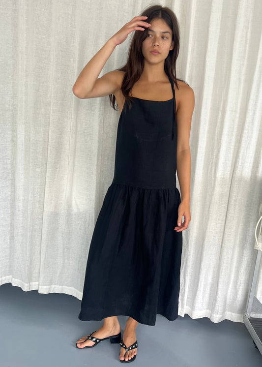 A woman stands indoors before white curtains, wearing the Gathered Halter Dress - Black by Deiji Studios. The sleeveless dress features a square neckline and ruffled hem. She is barefoot, touching her forehead with a neutral expression.