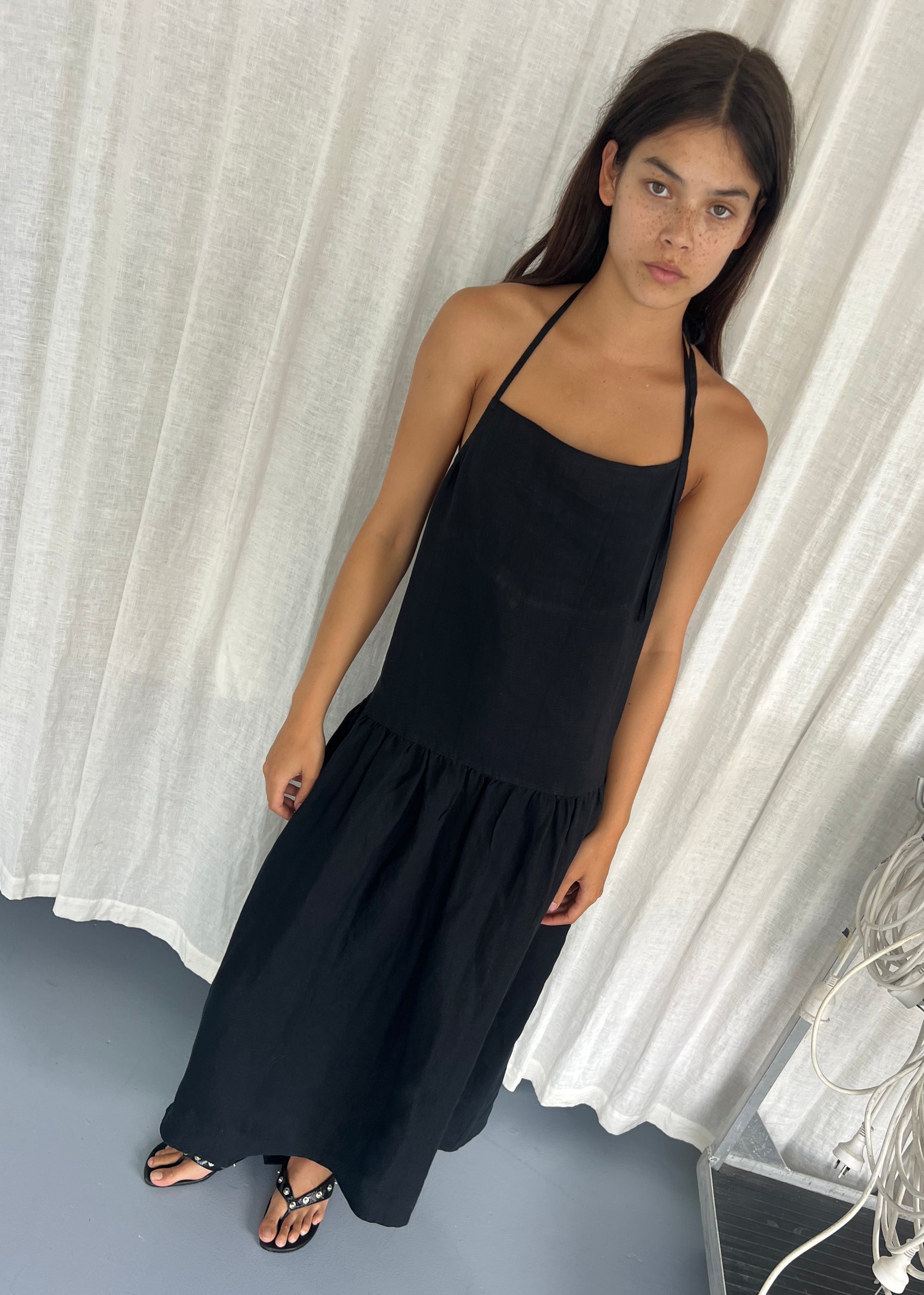 A young woman with long brown hair and freckles wears the Gathered Halter Dress - Black by Deiji Studios and black sandals, standing indoors on a gray floor in front of white curtains.