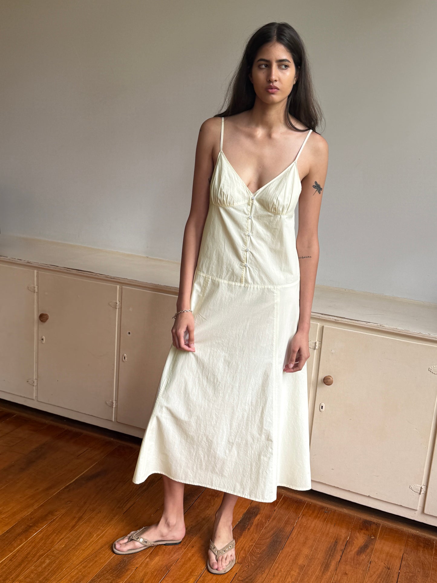 A woman with long dark hair and a small upper left arm tattoo stands indoors on a wooden floor before cream cabinets, wearing the Deiji Studios Double Seam Dress in Butter Yellow and sandals.