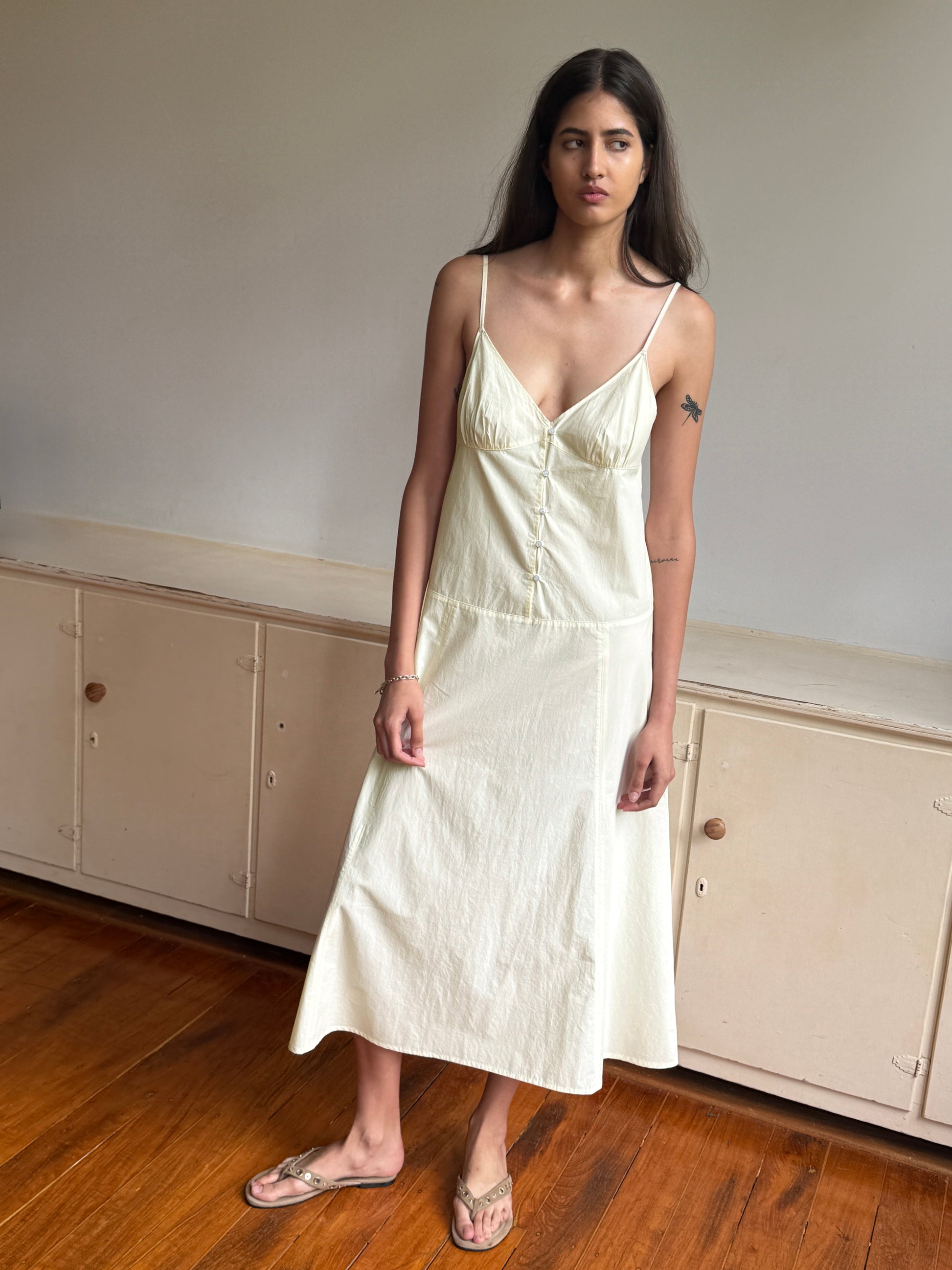 A woman with long dark hair and a small upper left arm tattoo stands indoors on a wooden floor before cream cabinets, wearing the Deiji Studios Double Seam Dress in Butter Yellow and sandals.