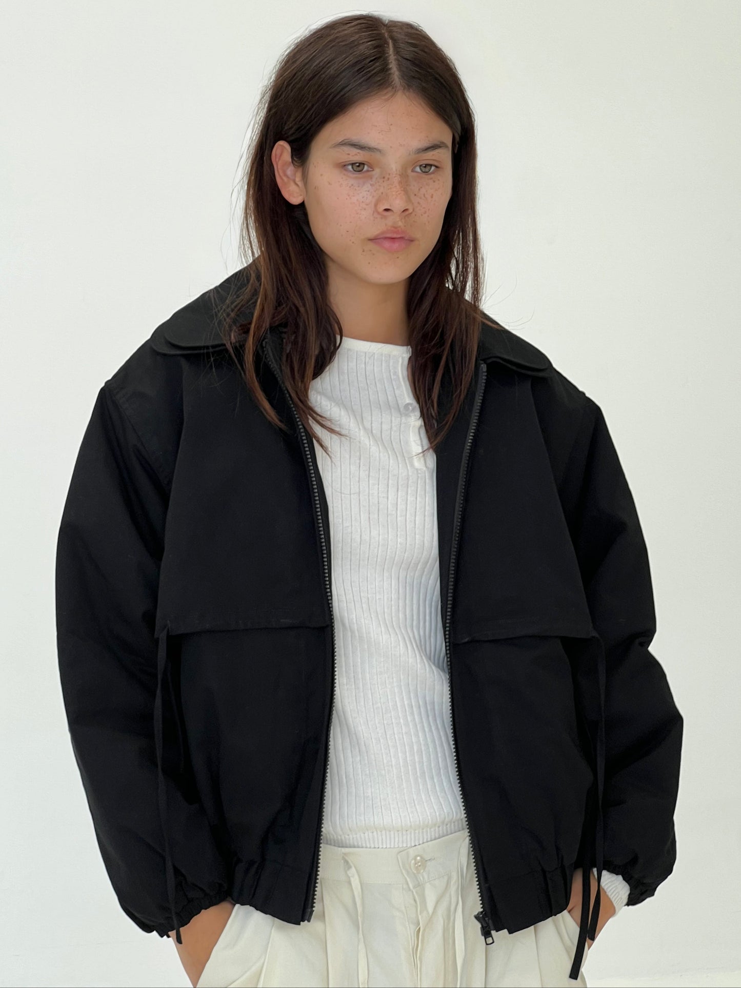 A young woman with straight brown hair and freckles wears the Double Layered Jacket - Black from Deiji Studios over a white ribbed shirt and light-colored pants, standing against a plain white background with a neutral expression.