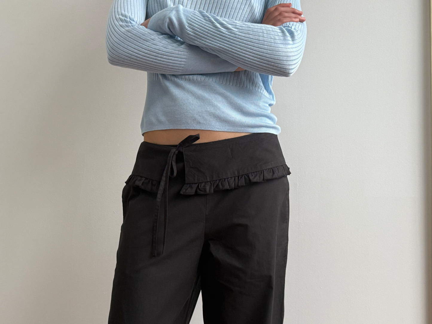 A person stands against a plain light-colored wall, arms crossed, wearing a light blue ribbed long-sleeve top and the Flounce Belt Pant - Black by Deiji Studios, featuring a ruffled waistband and tied belt.