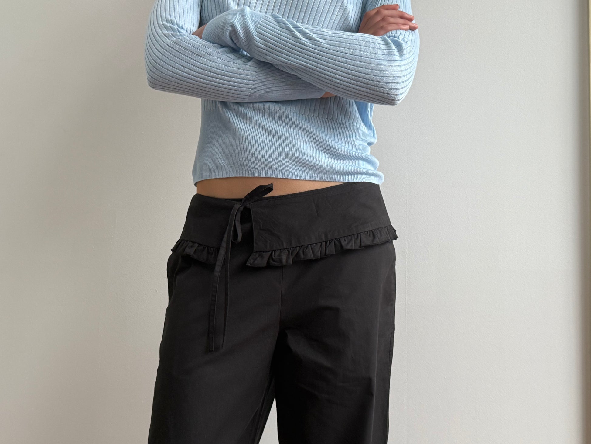 A person stands against a plain light-colored wall, arms crossed, wearing a light blue ribbed long-sleeve top and the Flounce Belt Pant - Black by Deiji Studios, featuring a ruffled waistband and tied belt.