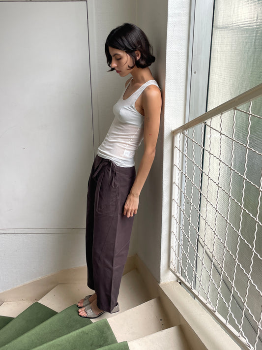 A woman with short dark hair stands on a staircase by a window, wearing a white tank top, Flag Tie Pant in Dark Brown, and sandals. She looks down and leans slightly against the wall.