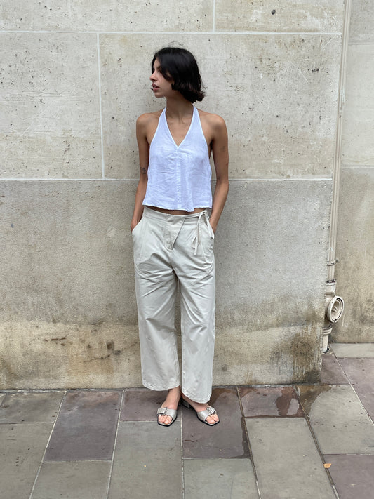 A person with short dark hair, wearing a sleeveless white halter top and Flag Tie Pant in Off White, stands against a weathered stone wall with hands in pockets, looking to the side.