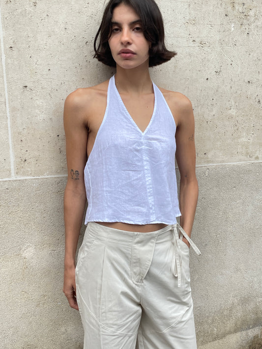 A person with short dark hair stands against a beige wall, wearing a sleeveless white halter top and the Flag Tie Pant - Off White, featuring a relaxed straight leg and side flag tie detail, looking slightly to the side with a neutral expression.