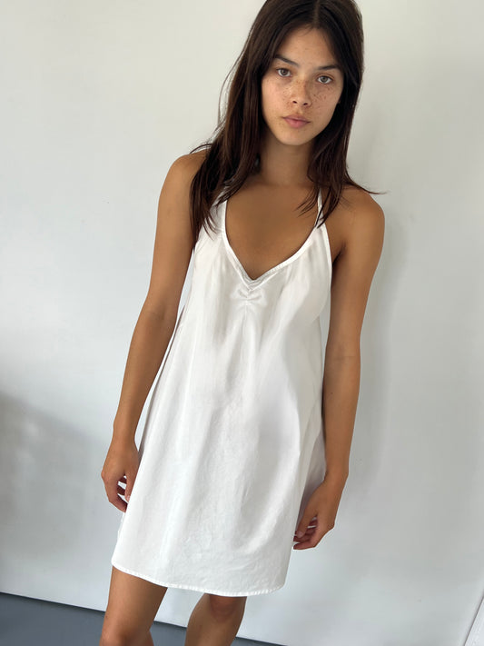 A young woman with long dark hair and freckles stands against a plain white wall, wearing the Round Halter Dress - White. She looks directly at the camera with a neutral expression.