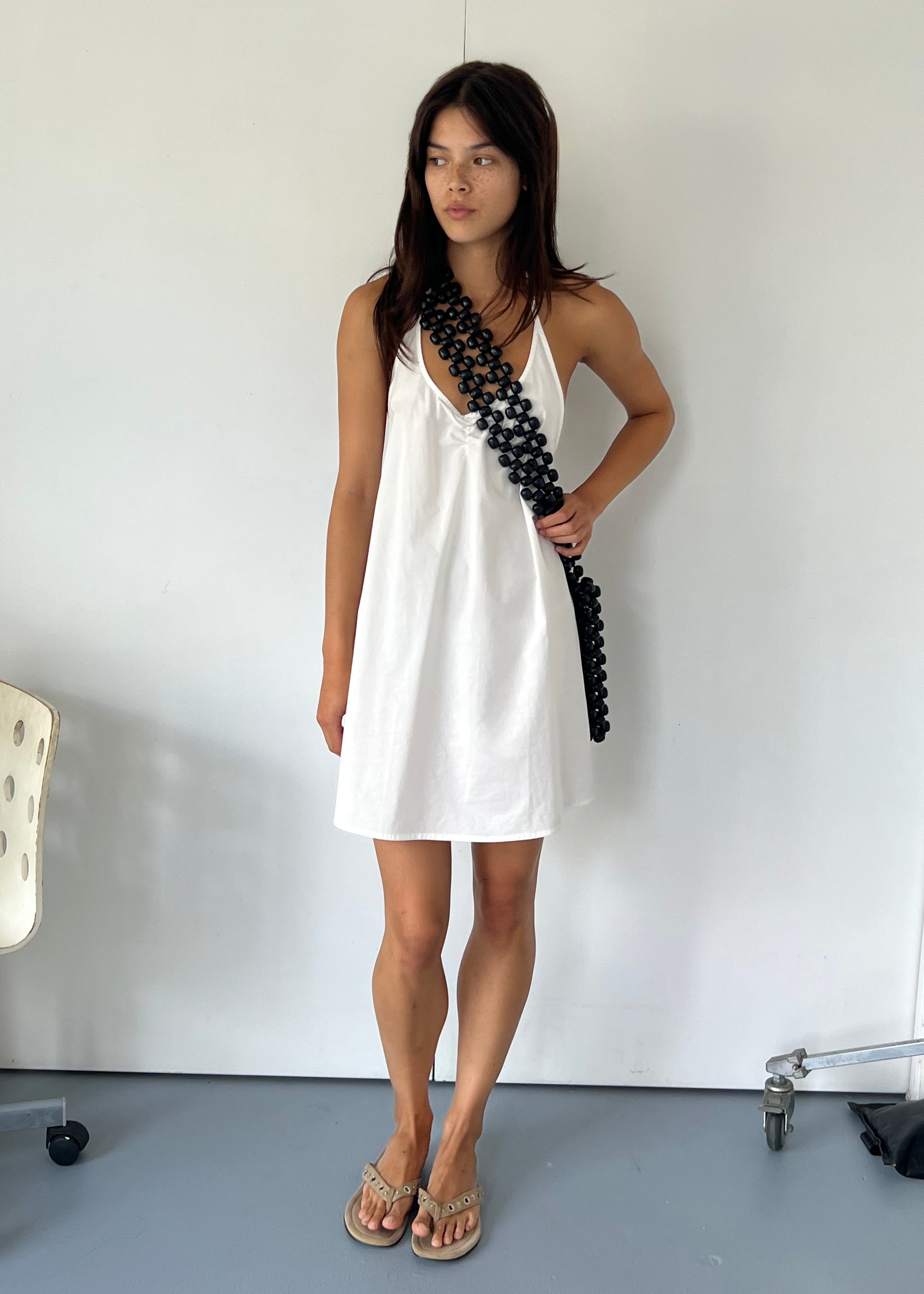A woman stands indoors against a white wall, wearing the Round Halter Dress - White by Deiji Studios and tan sandals. She has straight dark hair and a chunky black chain strap slung diagonally across her body.