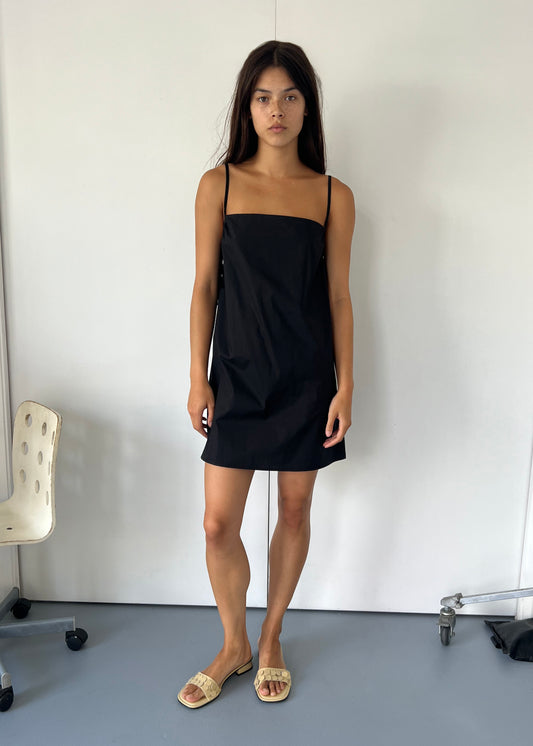 A woman with long dark hair stands indoors against a plain white wall, wearing the Drawcurve Dress - Black and light sandals. Nearby are an empty chair and a metal object on wheels.