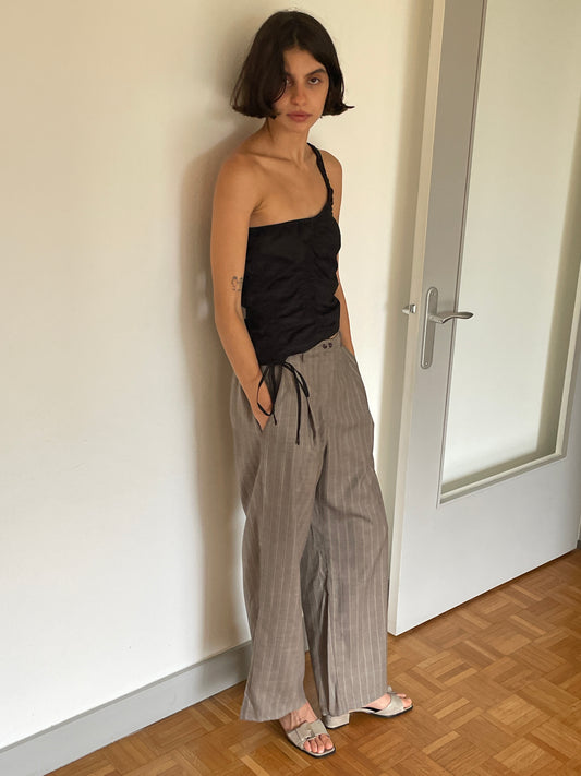 A woman with short dark hair stands indoors by a door, wearing the black Scrunchie Top from Deiji Studios, gray pinstripe wide-leg pants, and sandals. She looks at the camera with a neutral expression.