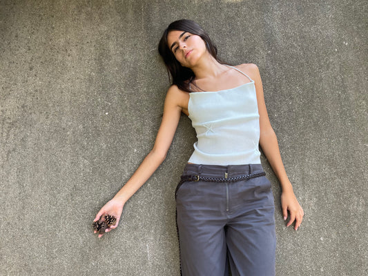 A woman with long dark hair lies on concrete, wearing a Deiji Studios Fine Knot Tank in Ice Blue and gray pants. Holding a small pine cone, she gazes upward with a relaxed expression.