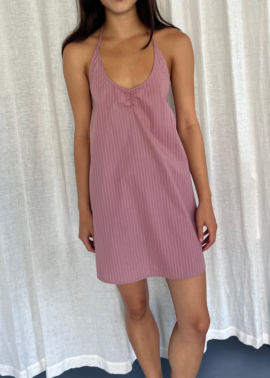 A woman stands before white curtains, wearing Deiji Studios' Round Halter Dress - Salt Stripe in pink with subtle vertical stripes. Her head is not visible, and her arms rest by her sides.