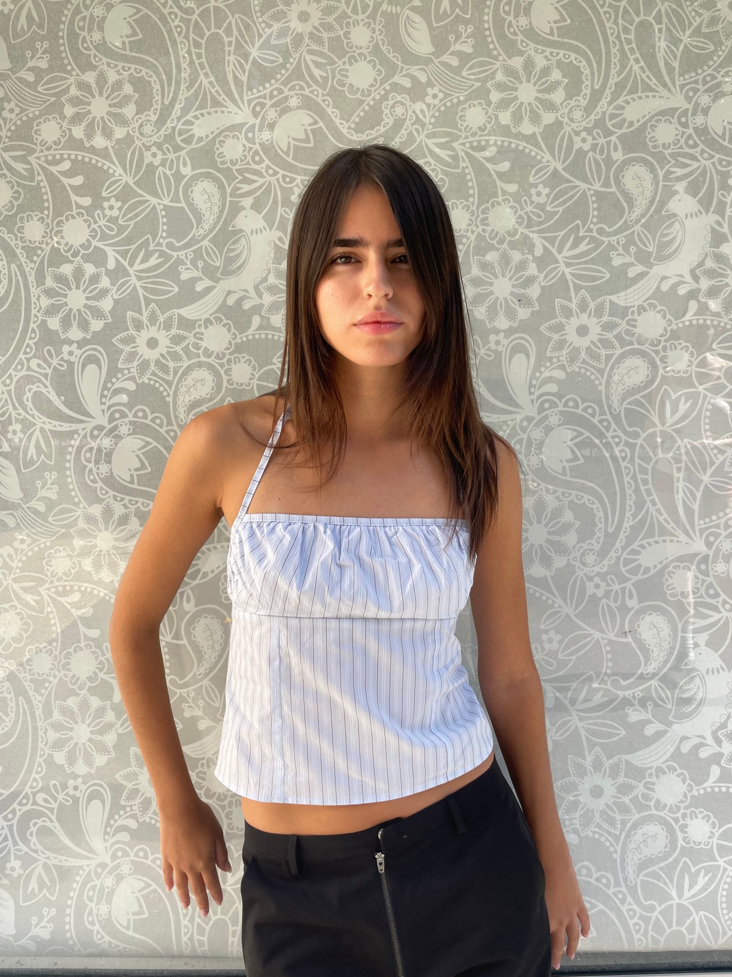 A young woman with long brown hair stands against a light floral background, wearing the Strap Top - Mineral Stripe, an organic cotton blue and white striped top, paired with black pants and looking at the camera neutrally.