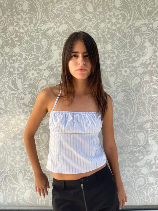 A young woman with long brown hair stands against a light floral background, wearing the Strap Top - Mineral Stripe, an organic cotton blue and white striped top, paired with black pants and looking at the camera neutrally.