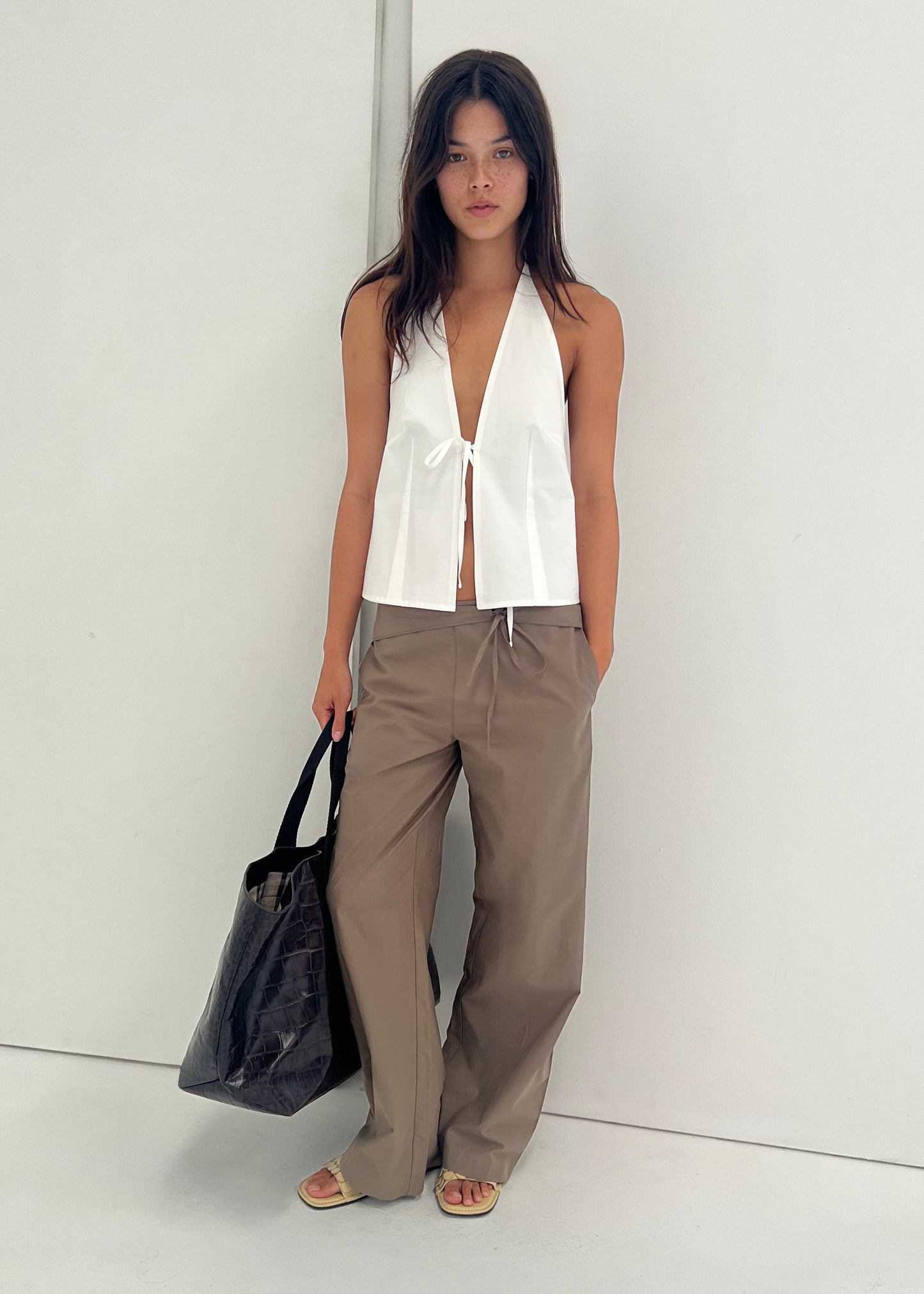 A woman stands against a white wall in a sleeveless white halter top and loose Deiji Studios Belted Pant - Clay. She holds a large black tote, wears sandals, and has her hands in her pockets with a relaxed expression.