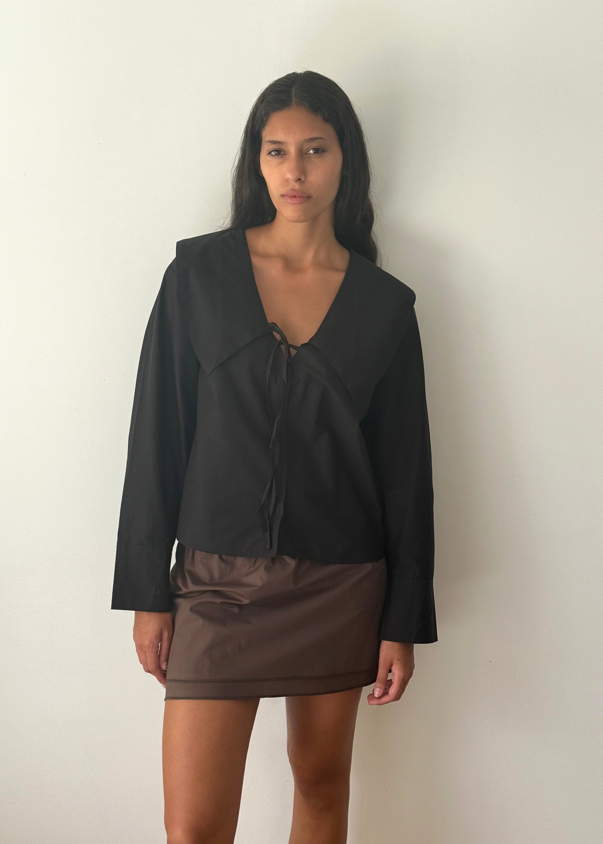 A woman with long dark hair and a neutral expression stands against a plain white wall, wearing the Collared Tie Shirt - Black with a wide collar and front tie, paired with a short brown skirt.