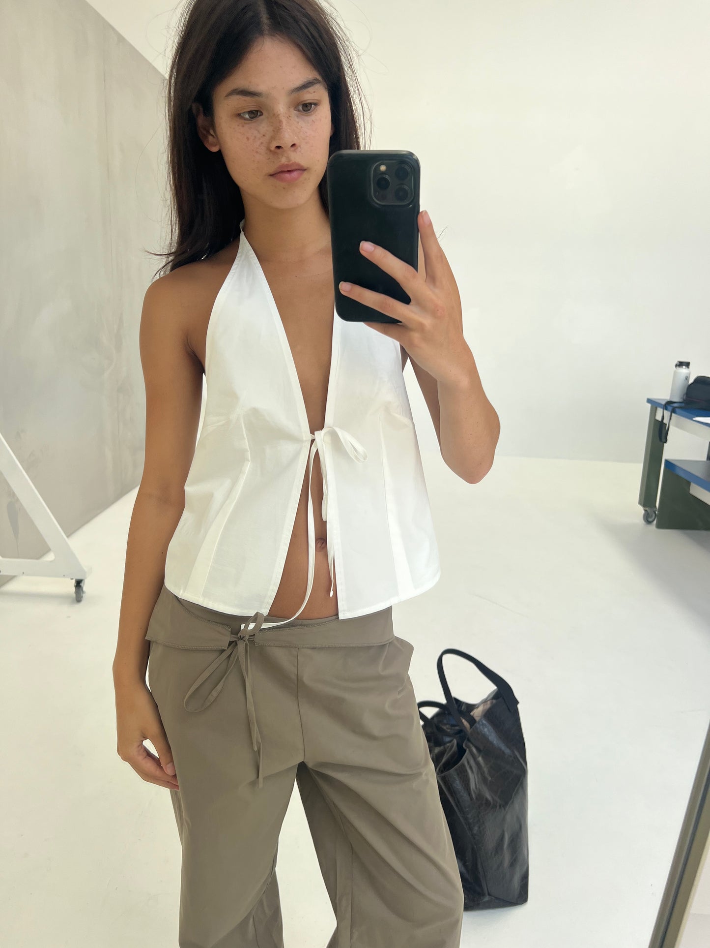 A person with long dark hair takes a mirror selfie in a minimal, light-filled room. They wear a sleeveless white halter top and the Belted Pant - Clay, with a large black bag on the floor beside them.
