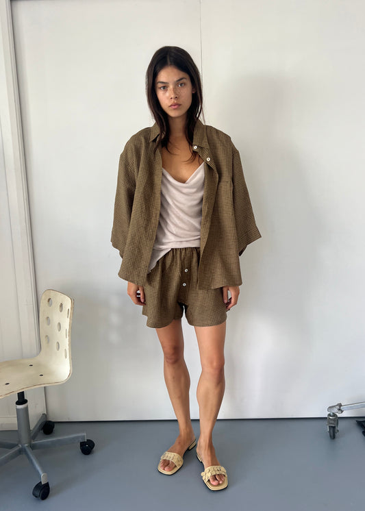 A woman with long dark hair wears the 03 Set - Pine Check: an oversized linen shirt and matching boxer-style shorts over a beige tank, paired with sandals. She stands indoors near a white chair against a plain wall.