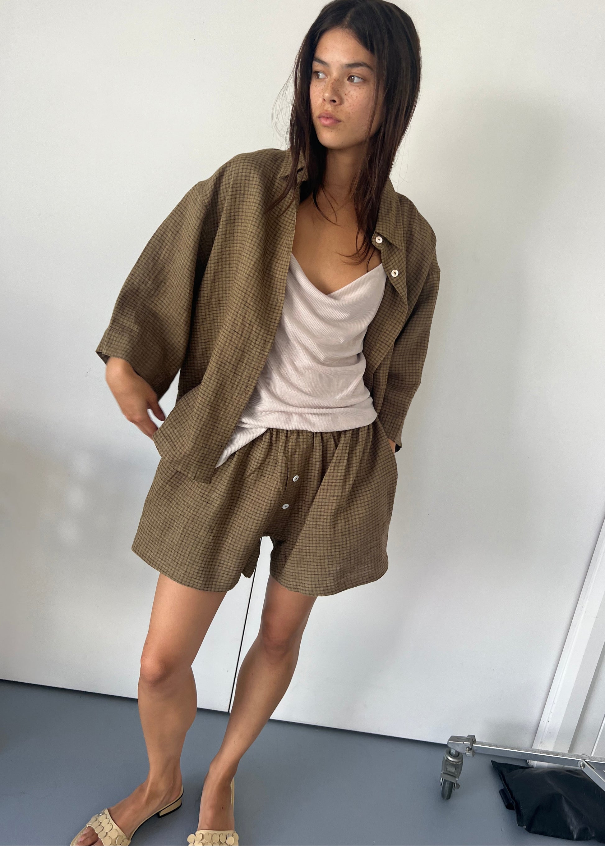 A woman with long dark hair stands indoors, wearing the 03 Set - Pine Check: a light beige tank top, an organic French linen checked button-up shirt, and matching boxer shorts. She has one hand in her pocket and looks to the side.