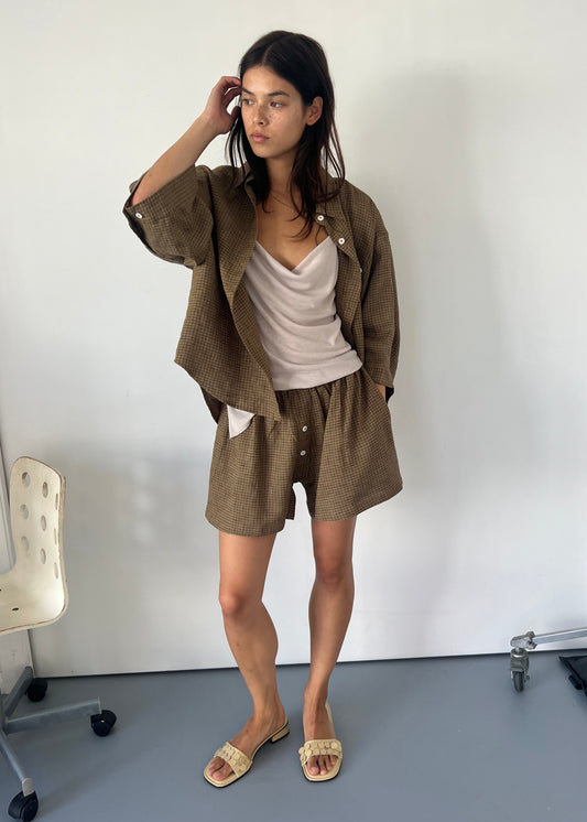 A woman with long dark hair stands indoors against a white wall, wearing the 03 Set - Pine Check: boxer-style shorts and an oversized brown check linen shirt, paired with a cream tank top and beige slide sandals, posing in a relaxed manner.