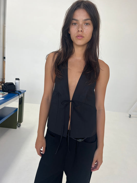 A woman with long brown hair wears the Open Halter Top - Black from Deiji Studios and black pants, standing indoors against a plain white background. She faces the camera with a neutral expression.