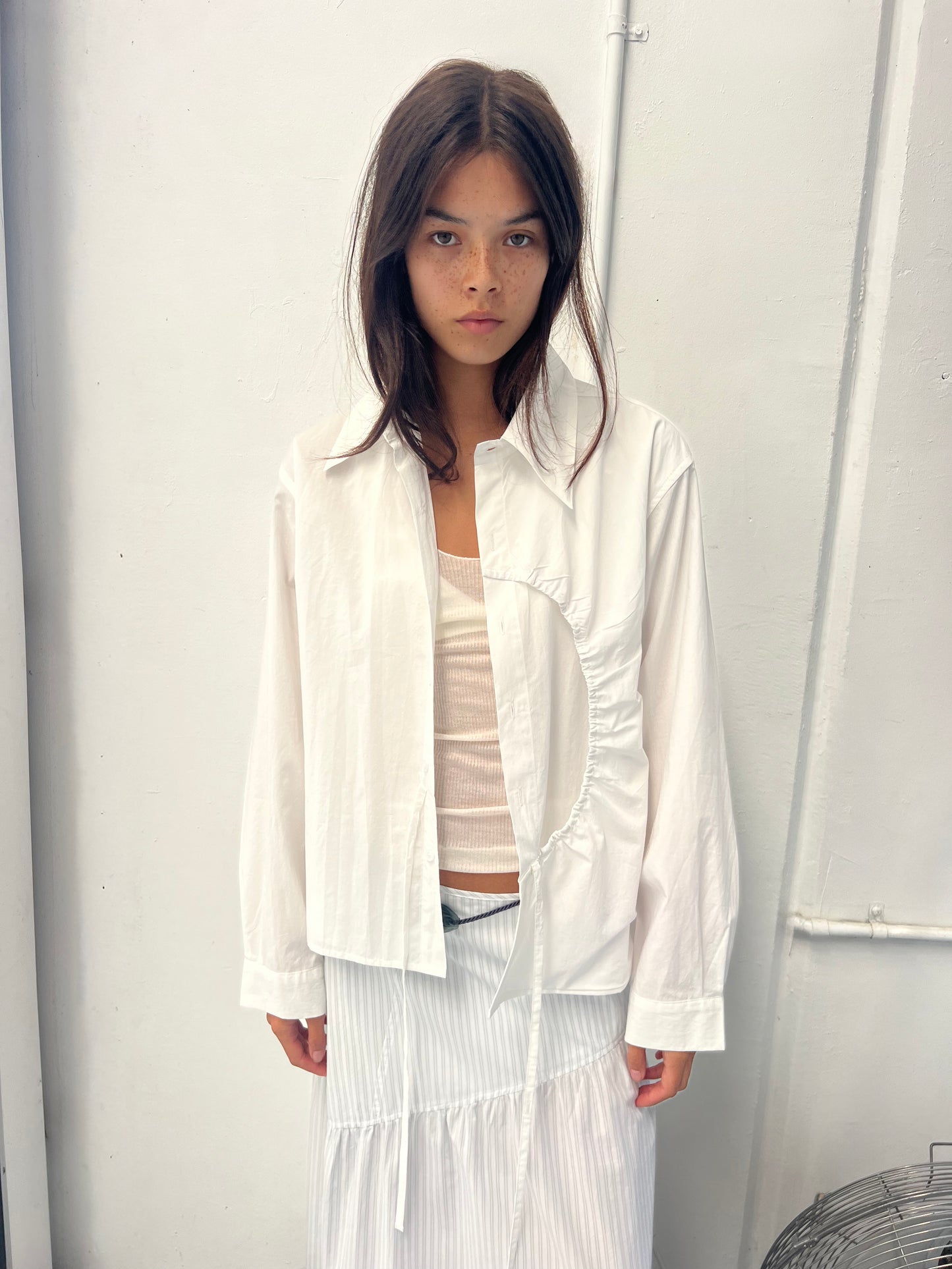 A woman with long dark hair stands against a white wall, wearing the Arch Tie Shirt - White by Deiji Studios in organic cotton poplin over a light top and pleated white skirt. The shirt features modern, asymmetrical details; she looks neutral.