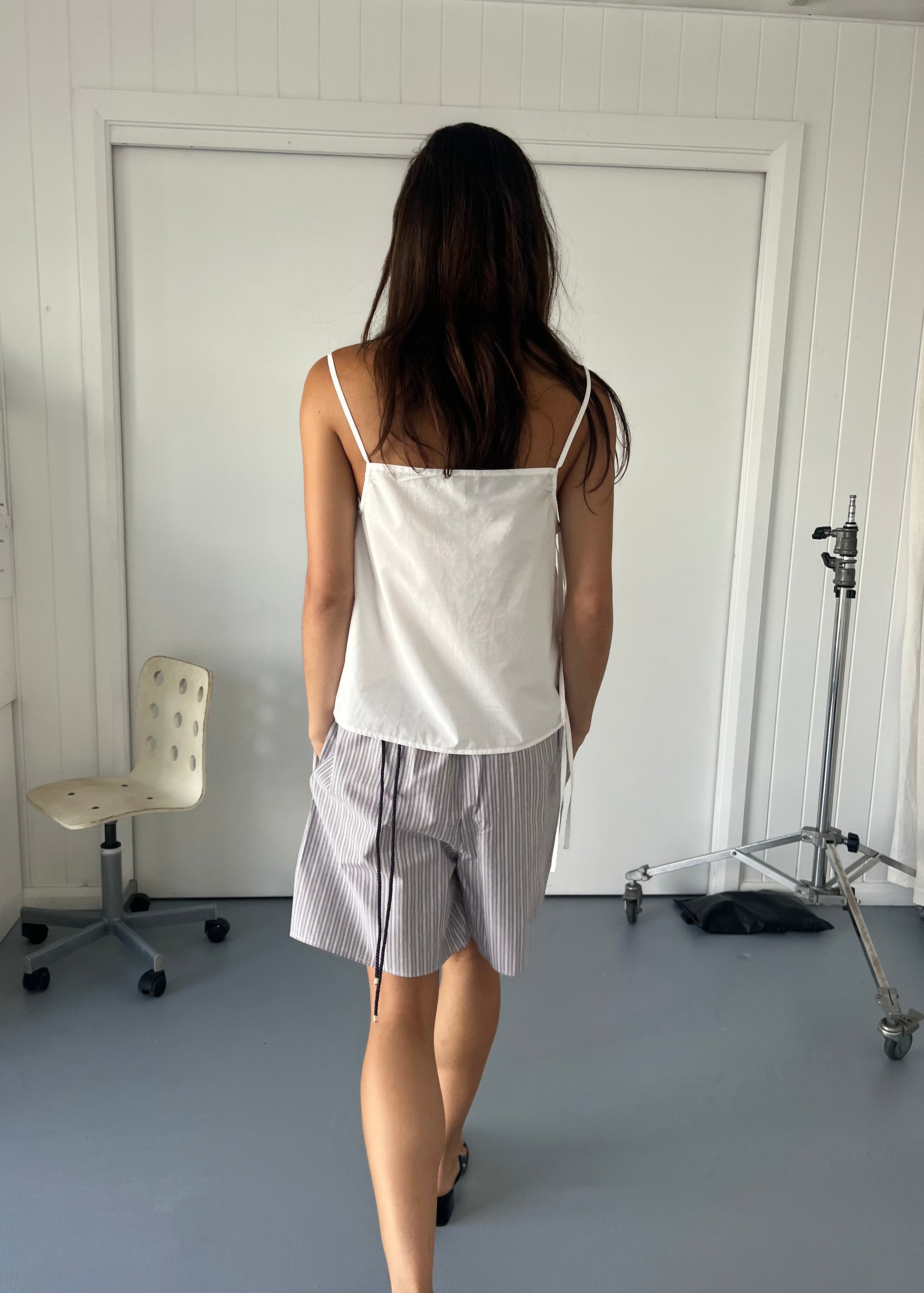 A woman with long dark hair stands indoors facing away, wearing a Deiji Studios Gathered Strap Top - White and gray striped shorts. An empty swivel chair and a metal stand are visible in the white-walled room.