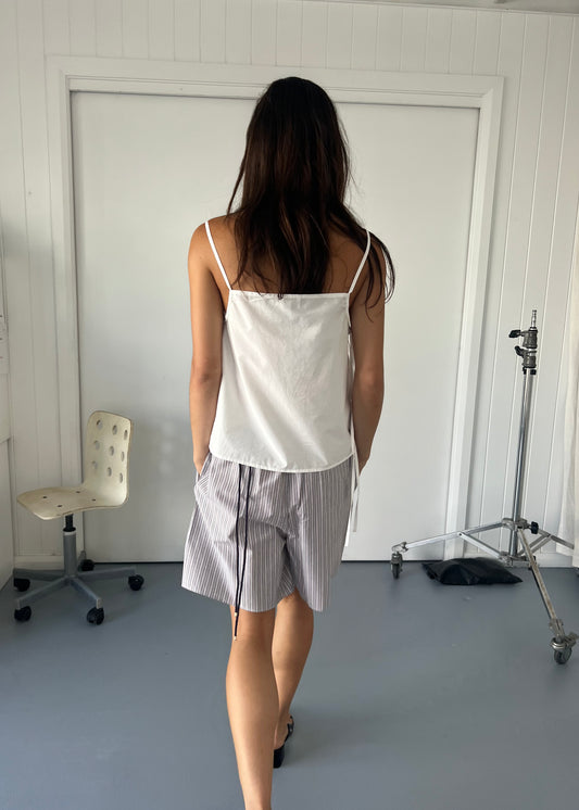 A woman with long dark hair stands indoors facing away, wearing a Deiji Studios Gathered Strap Top - White and gray striped shorts. An empty swivel chair and a metal stand are visible in the white-walled room.