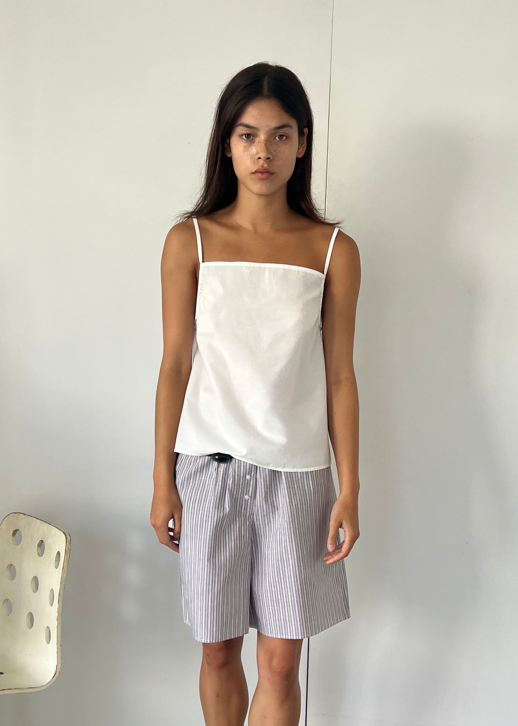 A young woman with long dark hair stands indoors against a plain white wall, wearing the Gathered Strap Top - White by Deiji Studios and light gray striped shorts. A white chair with circular cutouts is to her left.