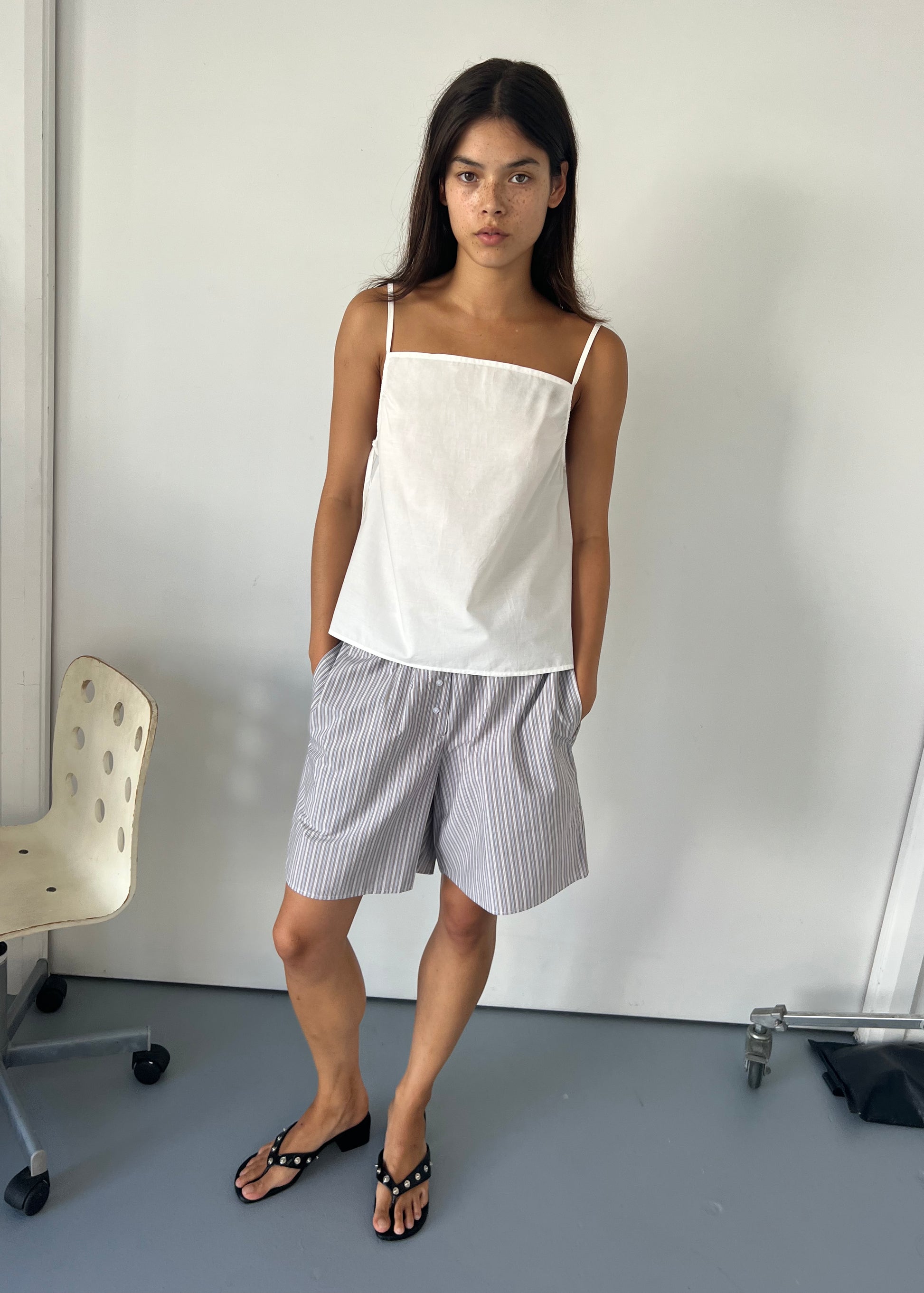 A woman with long dark hair stands indoors near a white chair, wearing the Deiji Studios Gathered Strap Top - White in organic cotton poplin, loose gray striped shorts, and black sandals. Her hands are in her pockets against a plain white wall.