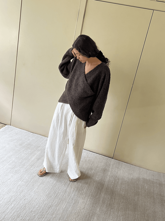 A person with long dark hair stands on a light rug, wearing the Crossover Top - Brown Multi by Deiji Studios with wide white pants and sandals, looking down and touching their hair against a neutral wall background.