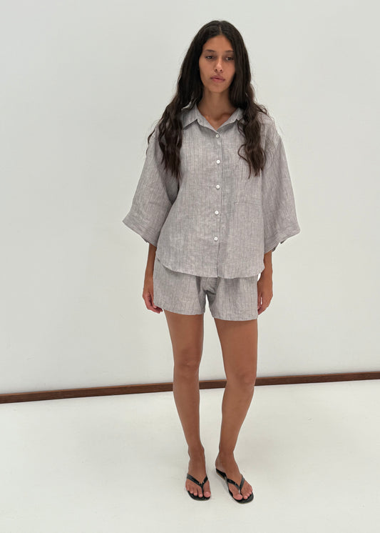 A woman stands indoors against a plain white wall, wearing the 03 Set - Haze Blue: a loose-fitting organic French linen shirt and matching boxer shorts, paired with black flip-flops.