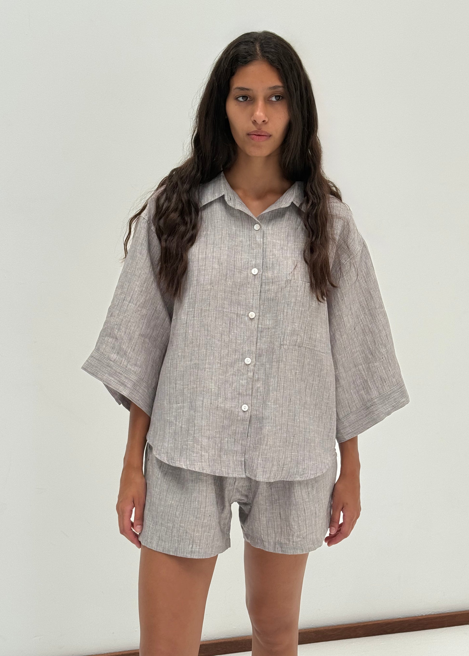 A woman with long wavy hair wears the 03 Set in Haze Blue—an organic French linen shirt with wide sleeves and matching boxer shorts—standing against a plain white background.
