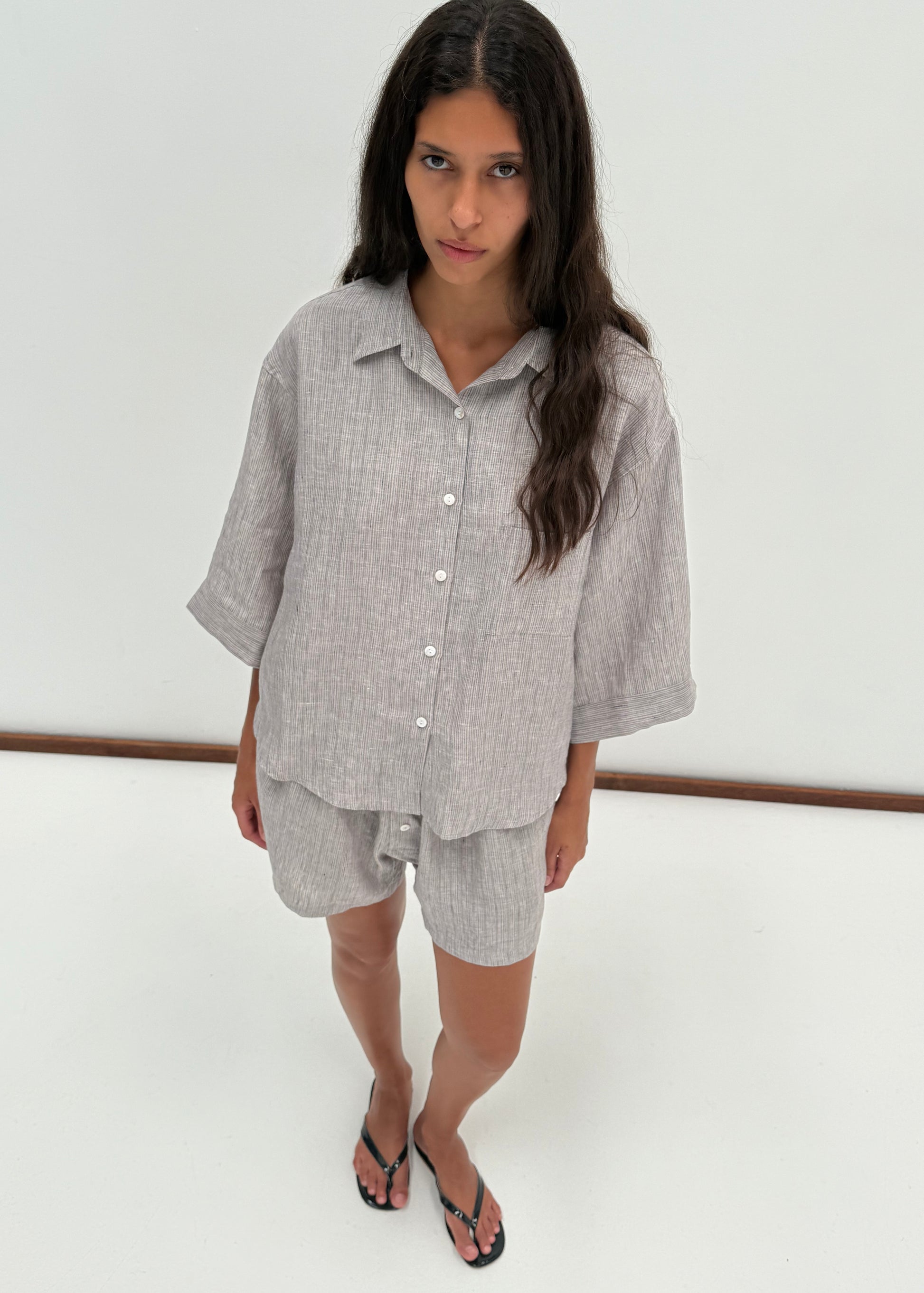 A woman with long dark hair models the 03 Set - Haze Blue, featuring a loose shirt and matching boxer shorts, with black flip-flops. She stands on a white floor against a plain white background, looking slightly up at the camera.