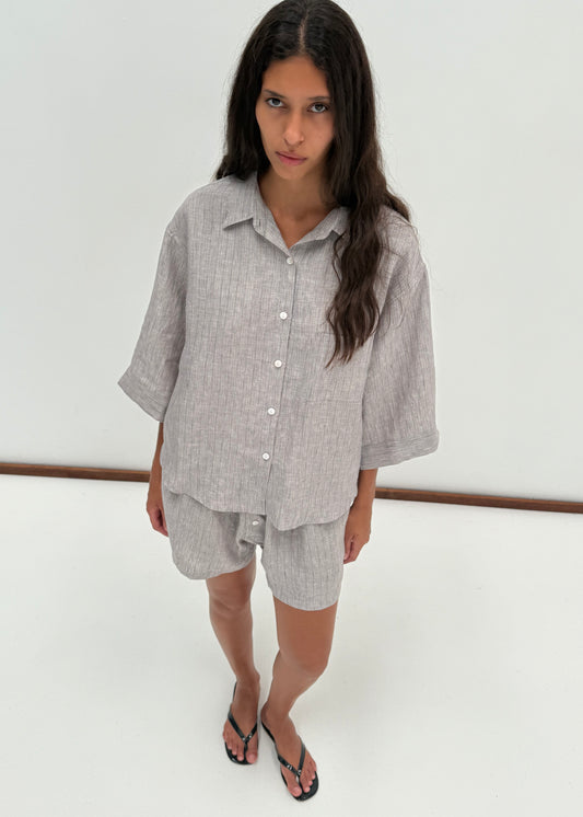 A woman with long dark hair models the 03 Set - Haze Blue, featuring a loose shirt and matching boxer shorts, with black flip-flops. She stands on a white floor against a plain white background, looking slightly up at the camera.