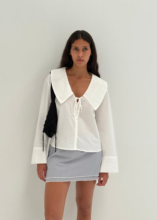 A woman with long dark hair and a neutral expression stands against a plain white wall, wearing the Collared Tie Shirt - White, a light blue and white striped mini skirt, and carrying a small black woven shoulder bag.