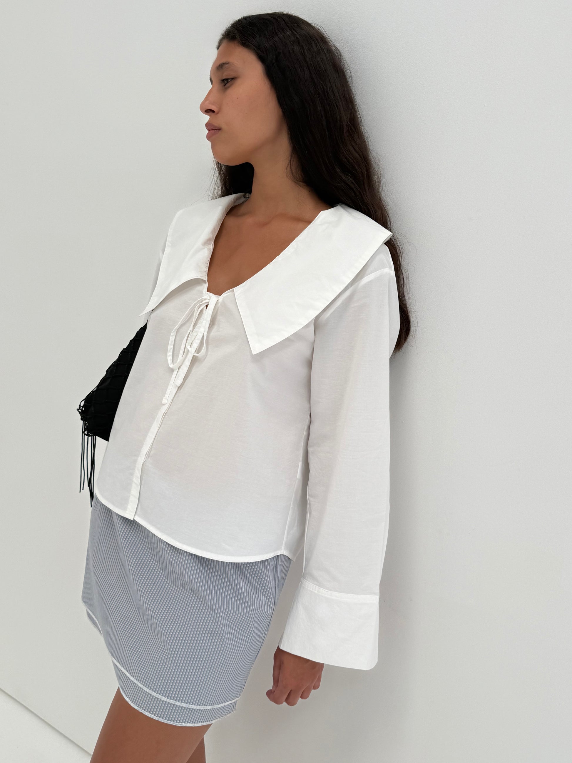 A woman with long dark hair stands against a white wall, wearing the Collared Tie Shirt - White with a large collar and paired with a blue and white striped skirt. She looks to the side, maintaining a neutral expression.