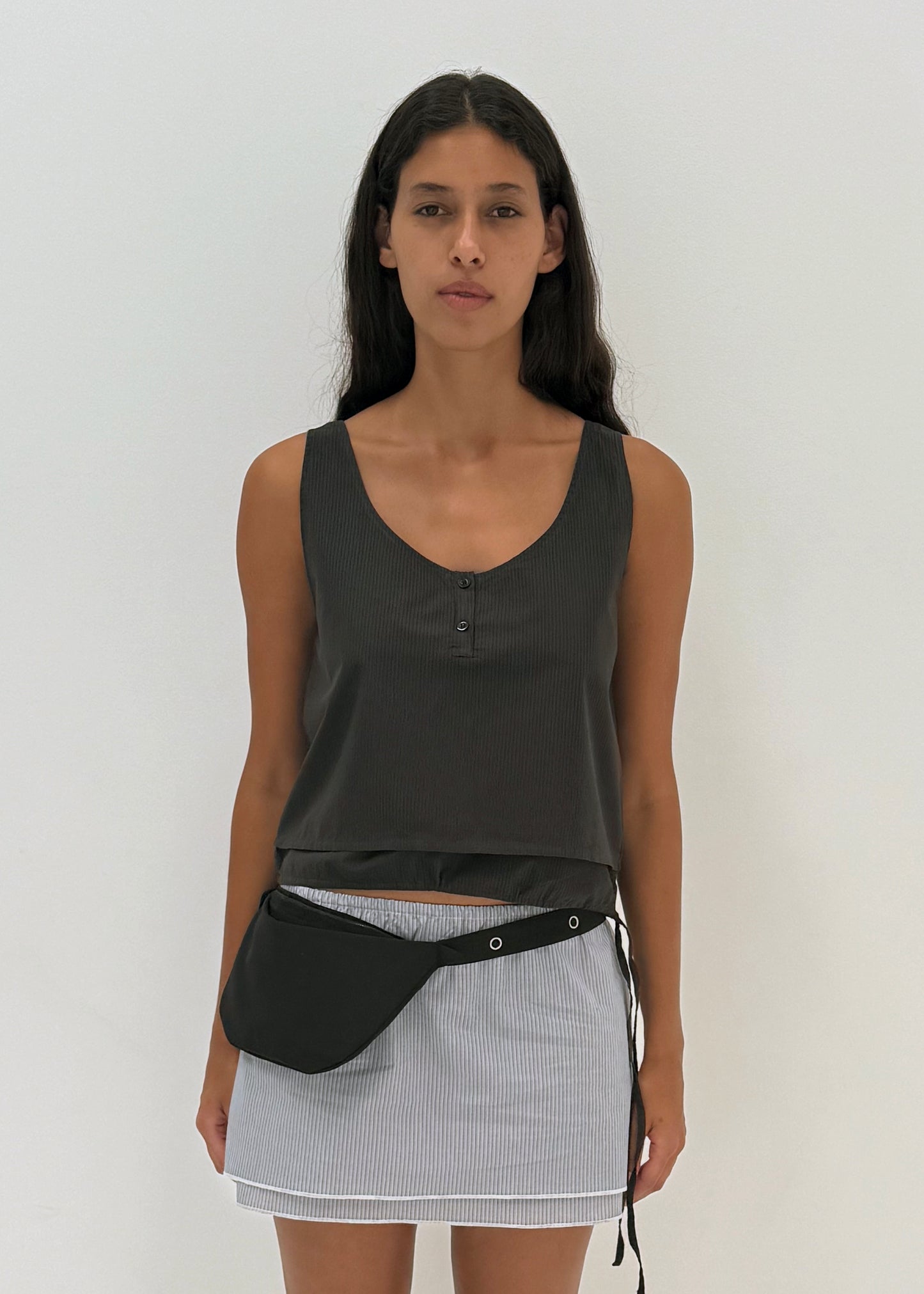 A woman with long dark hair wears the Layered Top - Black Stripe by Deiji Studios, paired with a light striped skirt and a black waist bag, standing against a plain white background.