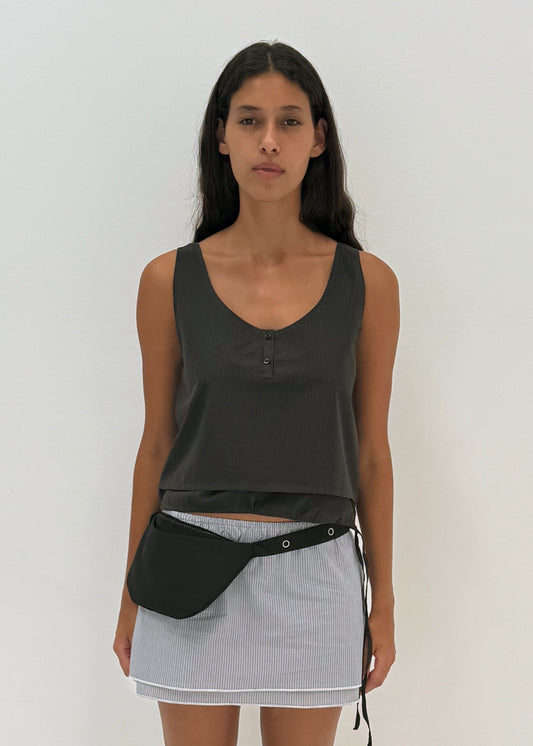 A woman with long dark hair wears a Deiji Studios Layered Top - Black Stripe, a short light gray skirt, and a black waist bag, standing against a plain white background.