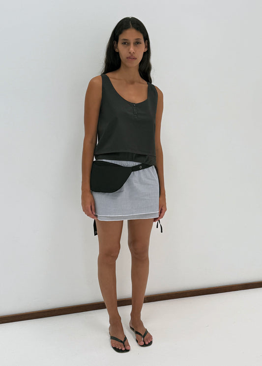 A woman with long dark hair and a neutral expression stands against a plain white wall, wearing the Layered Top - Black Stripe by Deiji Studios, a light mini skirt with white hem, a black waist bag, and flip-flops.