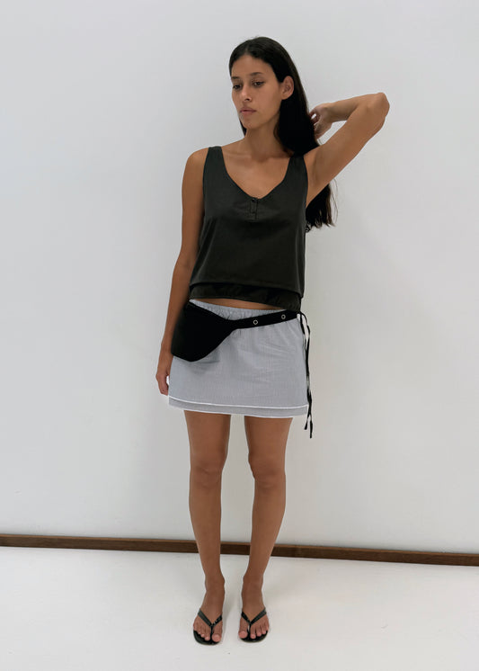 A woman with long dark hair stands against a white wall wearing the Layered Top - Black Stripe, a light mini skirt with a black wrap, black flip-flops, and a black waist bag, looking slightly to the side.