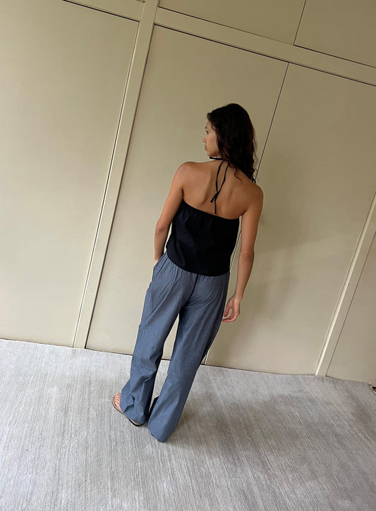 A woman with wavy brown hair stands facing away, wearing a black halter top and the Ease Trouser - Blue Pinstripe, relaxed with an elastic waist in organic cotton, paired with sandals on a light floor before beige paneled walls.
