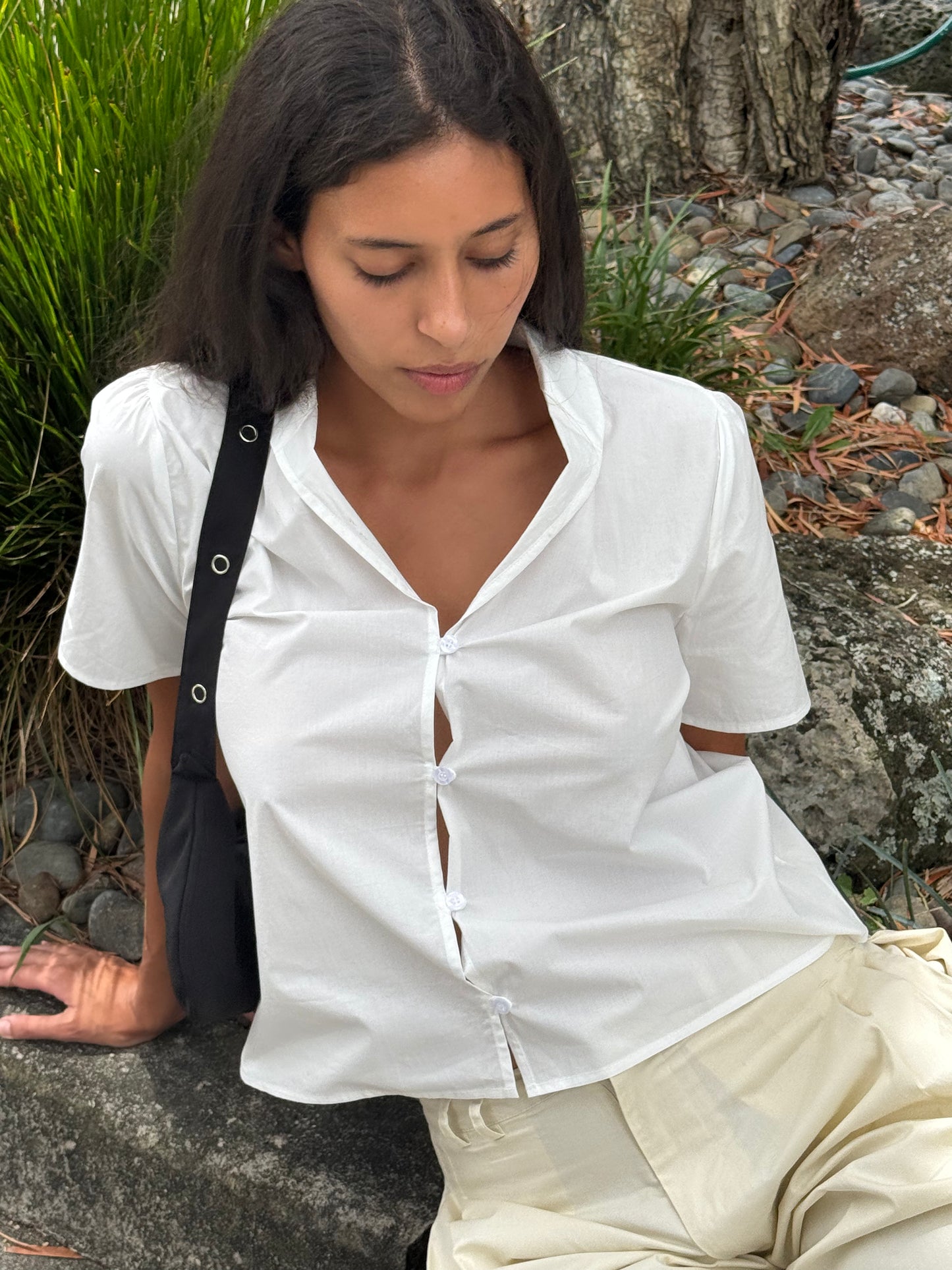 A woman with long dark hair sits outdoors on a rock, wearing the Sails Top in white and cream-colored pants, her black shoulder bag beside her. She looks down calmly, surrounded by green plants and rocks in the background.
