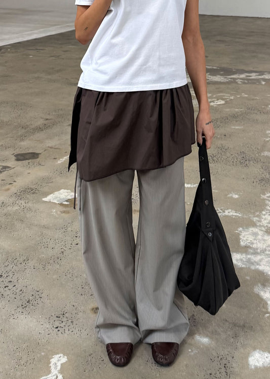 Wearing a white organic cotton t-shirt, loose dark brown tunic, wide gray pinstriped pants, and dark brown shoes, a person stands on a textured concrete floor holding a large black bag and sporting the Napkin Belt - Chocolate.